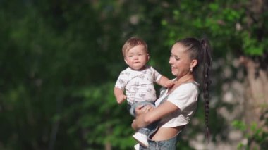 Gülümseyen anne tatlı çocuğunu kucağında tutuyor. Güzel anne ve küçük oğlu güzel göletin yanındaki parkta yürüyorlar. Tatlı çocuk annesinin kolyesiyle oynuyor. Annelik kavramı.