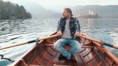 Hipster 'lı genç adam tatil boyunca Alph dağları manzarasının arka planındaki muhteşem gölün ortasındaki küçük teknede kürek çekiyor. Yakışıklı, gülümseyen adam harika manzaranın tadını çıkarıyor..