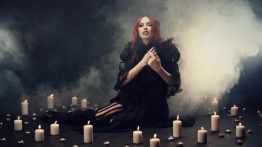 Kızıl saçlı cadı duman içinde oturmuş şeytanları çağırmak için enerjik el hareketleri yapıyor. Şeytan 'ı çağırmak için büyülü bir ayin. Cosplay, cadılar bayramı kostümü. Doğaüstü, kötü, karanlık, gotik kavramı.