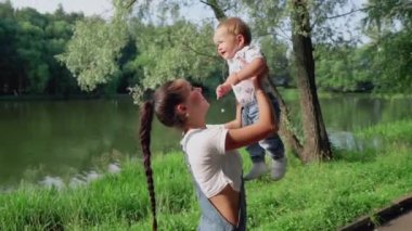 Güzel anne ve küçük oğlu güzel göletin yanındaki parkta yürüyorlar. Gülümseyen anne tatlı çocuğunu fırlatıyor. Birlikte mutlu olmak. Annelik kavramı, mutluluk, aşk ve güzellik..