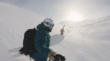 Snowboardcuları selfie çubuğuyla çeken adam. Rüzgar yükselen karla yokuş yukarı yürüyoruz. Güneşli bir günde dağların ve mavi gökyüzünün muhteşem arka planı. Keşif, seyahat, spor, hava durumu.