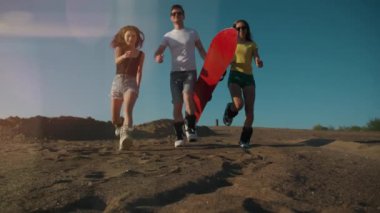 Yaz yaşam tarzı. Kırmızı snowboardlu üç genç koşuyor ve kameraya kum atıyor. Tasasız gençler birlikte eğleniyorlar. Özgürlük, spor, ekstrem, kum kaykayı, seyahat, turizm, tatil konsepti.