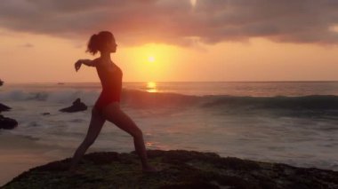 Mayo giymiş güzel bir kadın okyanus kıyısında yoga yapıyor. Dalgalar kuma vururken egzersiz yapan genç bir kızın silueti. Kadın güneş ışığı önünde meditasyon yapıyor. Sağlık kavramı, rahatlama, yoga.