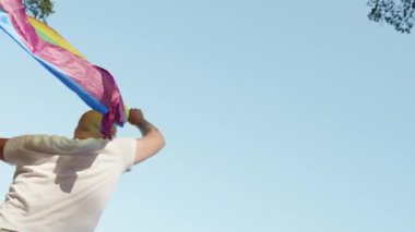 Mavi gökyüzünün arka planında geçit töreni yaparken elinde gökkuşağı eşcinseli bayrağı tutan eşcinsel adam. Kalp gözlüğü takan mutlu adam haklarını gösteriyor. LGBTQI, Gurur Etkinliği, LGBT Gurur Ayı, Eşcinsel Onur Sembolü