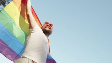 Mavi gökyüzünün arka planında geçit töreni yaparken elinde gökkuşağı gay bayrağı tutan eşcinsel bir adam. Kalp gözlüğü takan mutlu adam haklarını gösteriyor. LGBTQI, Gurur Etkinliği, LGBT Gurur Ayı, Eşcinsel Onur Sembolü