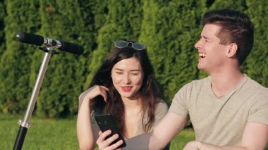 Erkek ve kız çimlerin üzerinde oturup ders çalışırlar ve akıllı telefon ekranına ya da cep telefonuna yakından bakarlar. Gençler ya da öğrenciler iletişim kurup gülüyor. Aktif yaşam tarzı, üniversite hayatı kavramı.