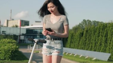 Güzel Asyalı kız akıllı telefondaki uygulamayla scooter 'ı kiralıyor. Kadın akıllı maaşla elektrikli scooter kiralıyor. Ekolojik toplu taşıma. Üniversite eko hayatı, aktif yaşam tarzı ve kiralama kavramı