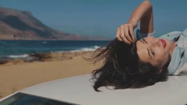 Arabanın çatısında kaygısızca yatan güzel genç bir kadın. Magnifficent Sahili 'nde arabanın yakınında. Sıcak yaz gününde saçlarına dokunarak hayatın tadını çıkaran bir kız. Seyahat, tatil, macera, gençlik, eğlence kavramı