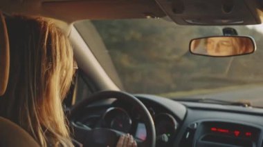 Güneş gözlüklü mutlu sarışının gün batımında arabasını sürerken müzik eşliğinde dans edişinin arka görüntüsü. Hoş bir kadın, müzik dinliyor ve gülümsüyor. Mutluluk ve neşe kavramı