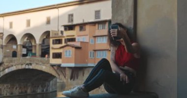 Yaz tatili sırasında Floransa 'ya seyahat eden genç bir kadının portresi. Kırmızı tişörtlü ve çizgili kot pantolonlu Asyalı kız Vecchio köprüsünde oturuyor ve akıllı telefonunda selfie çekiyor..