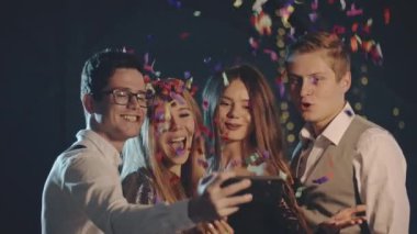 Gülümseyen çok ırklı arkadaşlar konfeti fırlatıp, akıllı telefondan veya cep telefonundan selfie çekiyorlar. Mutlu adamlar Noel partisinde modern teknolojiyi kullanarak eğleniyor ve fotoğraf çekiyorlar..