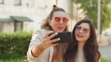 Olumlu duygular ve aşk. İki genç, gülümseyen hippi sarışın kadın akıllı telefondan kendi portresini çekiyor. Sokak geçmişinde çok şık kızlar var. LGBTQI, Gurur Etkinliği, LGBT Gurur Ayı