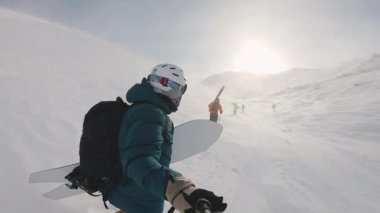 Snowboardcuları selfie çubuğuyla çeken adam. Rüzgar yükselen karla yokuş yukarı yürüyoruz. Güneşli bir günde dağların ve mavi gökyüzünün muhteşem arka planı. Keşif, seyahat, spor, hava durumu.