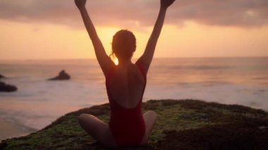 Yoga yapan genç bir kadının silueti. Okyanus kıyısında dinlenen kırmızı mayolu bir kız. Güzel esmer kadın, gün batımının önünde meditasyon yapıyor. Sağlık kavramı, rahatlama, yoga.