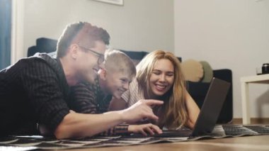 Genç mutlu ebeveynler ve yerde yatarken dizüstü bilgisayar kullanan sarışın çocuk. Gülümseyen aile bilgisayar ekranına bakıyor ve komik sosyal medya videoları izlemekten zevk alıyor. Evde olma kavramı.