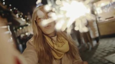 Yeni yıl arifesinde gülümseyen ve yanan parlak kıvılcımlar. Selfie zamanı, kız blogcu Prag Meydanı 'ndaki sokak fuarında sosyal sayfası için video ve fotoğraf çekiyor..