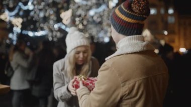 Kameraya karşı duran adam, eski kasaba meydanındaki Noel gecesinde kız arkadaşına bir hediye sunar. Aşk, aile, Noel, yeni yıl, tatil konsepti. RED kamera, 10 bit klonlama