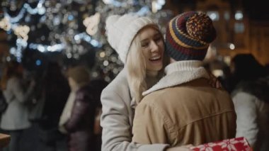 Kameraya karşı duran bir adam, eski kasaba meydanındaki Noel gecesinde kız arkadaşına bir hediye sunar. Aşk, aile, Noel, yeni yıl, tatil konsepti. RED kamera, 10 bit klonlama