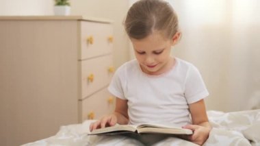 Sevimli küçük kız yatakta ders kitabı güneşli gün ile oturuyor. Çocuk okumayı öğrenme.