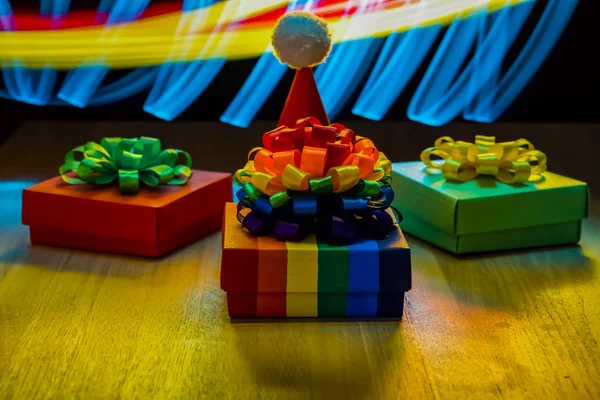 Christmas hat. Multicolored boxes with Christmas bows. Wooden table. Yellow, blue and red blurred stripes. Light effect close up.