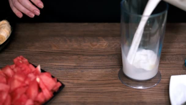 Cucina tavolo in legno. Versare il latte da piccolo in un bicchiere grande. Mettere nel latte i pezzetti di anguria matura tagliati a fette . — Video Stock