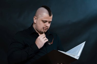 Serseri bir çalışma klasörü ile duruyor. Siyah gömlek. Iroquois. Gözlük çerçeve dudaklar dokunur. Mindfulness ve dalgınlık yüz ifadesi.