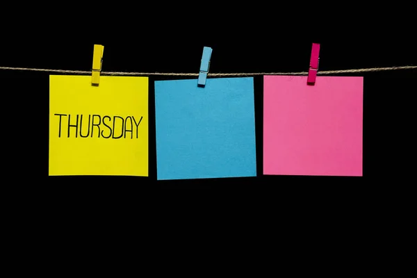 Kopierraum. Aufkleber auf dem schwarzen Hintergrund. Donnerstag. gelbe, rosa und azurblaue Papierblätter, die an einer Wäscheklammer befestigt sind. — Stockfoto