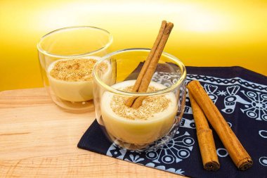 Üstünde tarçın olan iki bardak eggnog. Bir bardakta tarçın çubuğu var. Bardaklar ahşap bir kesme tahtasının üzerinde ve tatil desenli mavi bir kumaş..