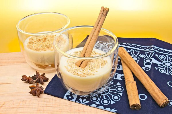 Стакан эггнога с корицей сверху и палочка корицы в одном из стаканов. Стекло сидит на рождественской синей ткани. Коричные палочки и звездный анис также на ткани. Все сидит на деревянной разделочной доске.