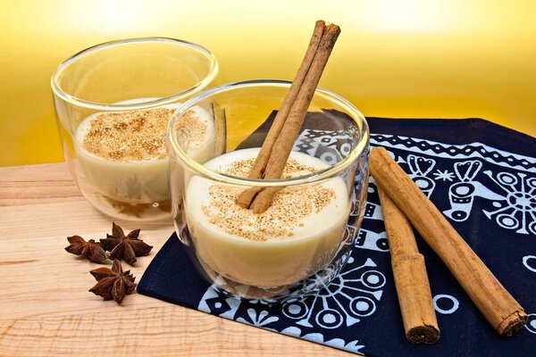 Две чашки эггнога с корицей в одном стакане и корицей в обоих стаканах. Звездный анис и коричные палочки сидят на разделочной доске и синей ткани с праздничными украшениями. На заднем плане металлическое золото.