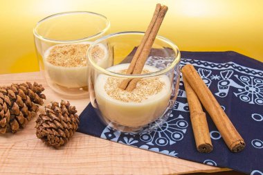 Bir bardakta tarçın çubuğu, iki bardakta da tarçın olan iki bardak eggnog. Çam kozalakları ve tarçın çubukları ağaç kesme tahtasına ve bayram süslemeleriyle mavi kumaşa otururlar. Arka plan metalik altından..