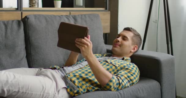 Young man is lying on a couch and uses his tablet — Stock Video