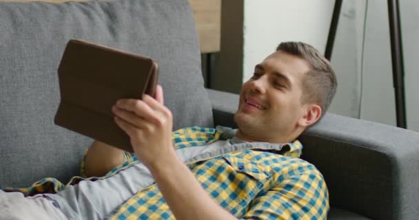 Hombre alegre se relaja en un sofá con su tableta — Vídeo de stock