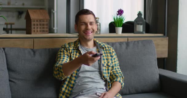 Un joven usa un mando a distancia, sentado en un sofá — Vídeos de Stock