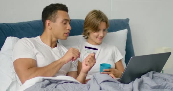 Joven pareja mixta está haciendo una compra en línea acostado en una cama — Vídeos de Stock
