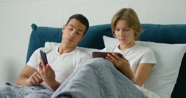 Молодая смешанная пара лежит в постели по утрам, используя гаджеты — стоковое видео