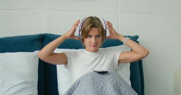 Young woman lying in bed, putting wireless headset on — Stock Video