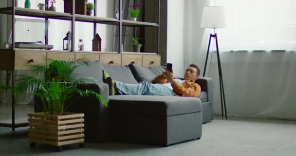 Homem hispânico está deitado no sofá, navegando no smartphone — Vídeo de Stock