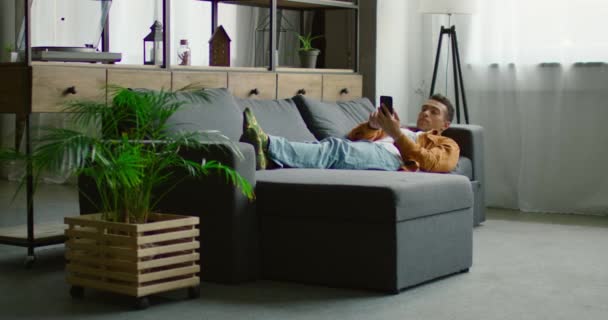 Homem hispânico está deitado no sofá, usando smartphone — Vídeo de Stock