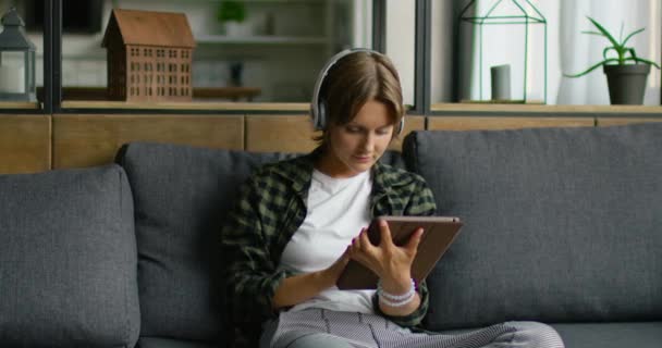 Kvinnan bär trådlöst headset, hålla Tablet PC, sitter på soffan — Stockvideo