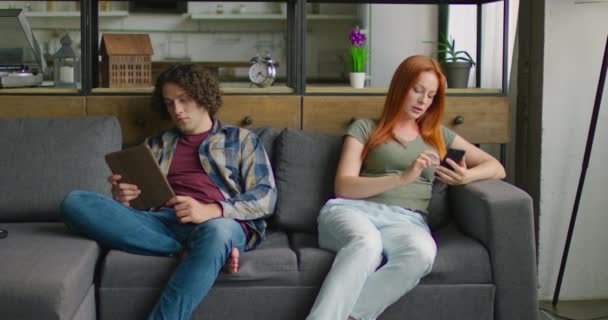 Giovane uomo e donna sono seduti sui bordi del divano, utilizzando i loro gadget — Video Stock