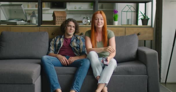 Молодая пара сидит на диване, женщина смотрит телевизор, мужчина скучает — стоковое видео