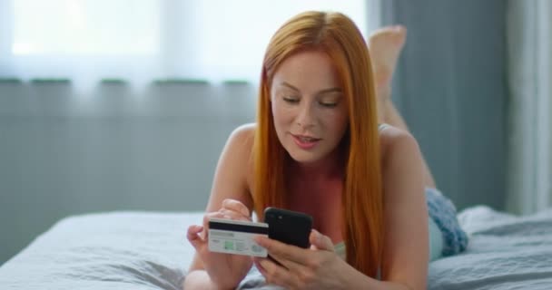 Hermosa mujer está acostada en la cama, ingresando el número de tarjeta en su gadget — Vídeos de Stock