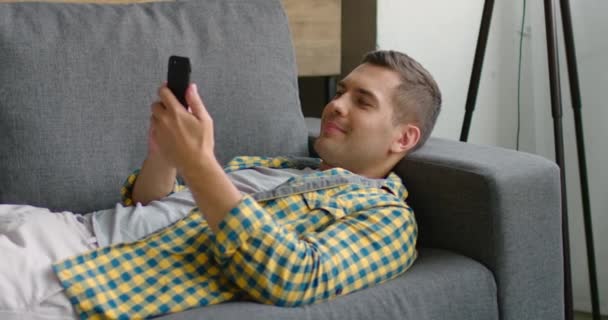 Jeune homme couché sur le canapé, en utilisant son gadget, textos, montage, navigation — Video