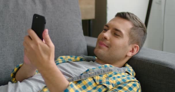 Jeune homme en tenue décontractée, allongé sur le canapé, utilisant son smartphone, gros plan — Video
