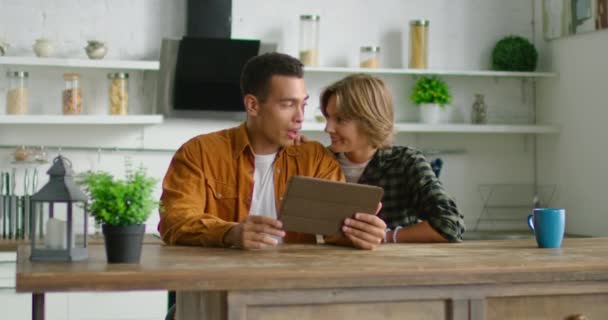 Boldog vegyes verseny pár ül a konyhában, segítségével tabletta számítógép — Stock videók