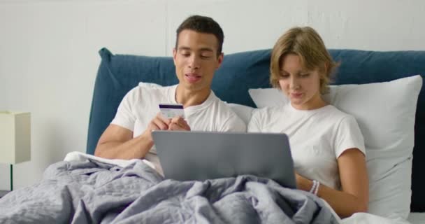 若い混合レースのカップルは、ベッドに横たわって、オンラインショッピングをしています — ストック動画