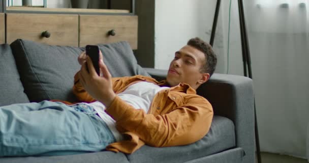 Красивий латиноамериканського людина розслабляє на дивані, сидячи у своєму смартфоні — стокове відео