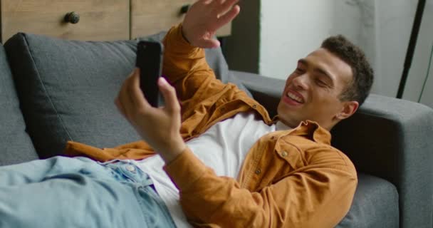 Hombre hispano está tumbado en el sofá, teniendo un chat de vídeo a través de teléfono inteligente — Vídeo de stock