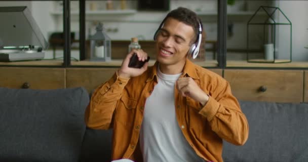 Pohledný latinský muž poslouchá hudbu, sedí na pohovce v obývacím pokoji — Stock video